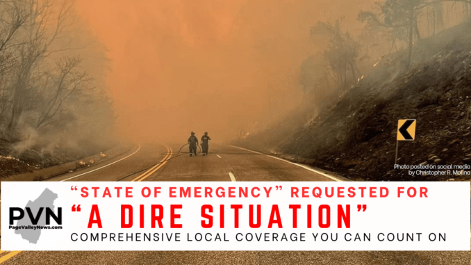 Two Luray Volunteer Firefighters with a hose on US 211 West of Luray. Either side of the roadway are scorched, with heavy smoke in the background. The text reads ""State of Emergency" requested for "a dire situation". Page Valley News graphic